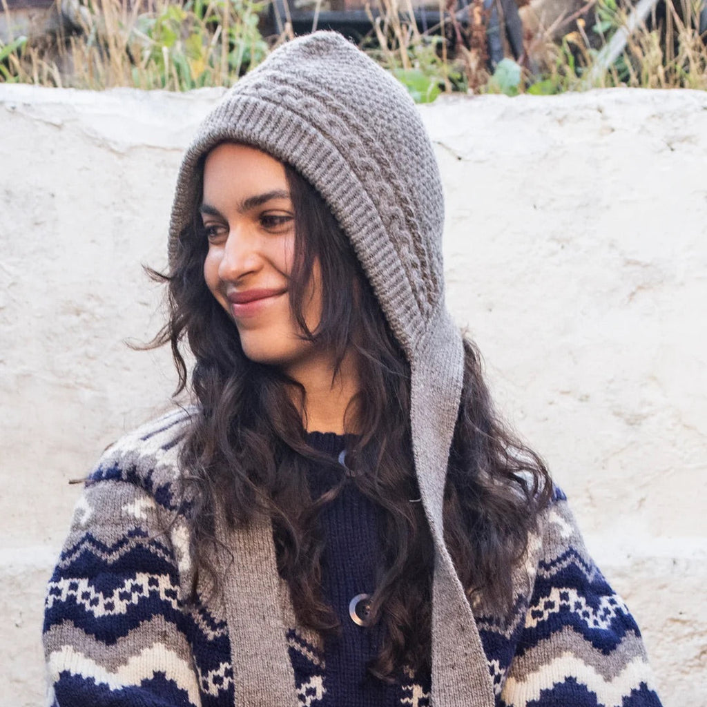 Woman wearing a patterned cardigan and knitted hat sitting on a stone wall.